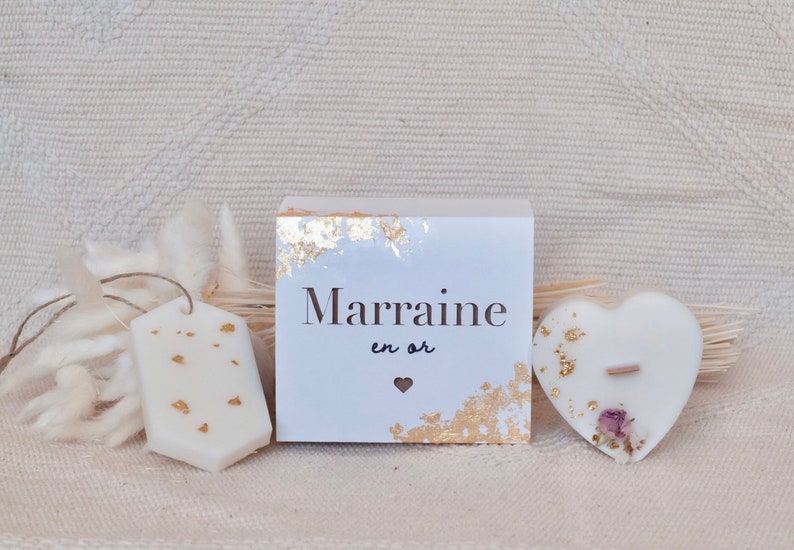 Op de afbeelding: Een verzameling witte kaarsen met goudblad accenten. Bevat een tag-vormige kaars, een doos met de tekst "Marraine en or", en een hartvormige kaars met een houten lont en gedroogde bloemen.