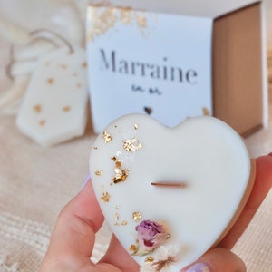 Op de afbeelding: Een hartvormige witte kaars met goudbladaccenten en gedroogde bloemen. De kaars heeft een houten lont en is versierd met kleine gedroogde bloemen. Een doos met het woord "Marraine" staat op de achtergrond.