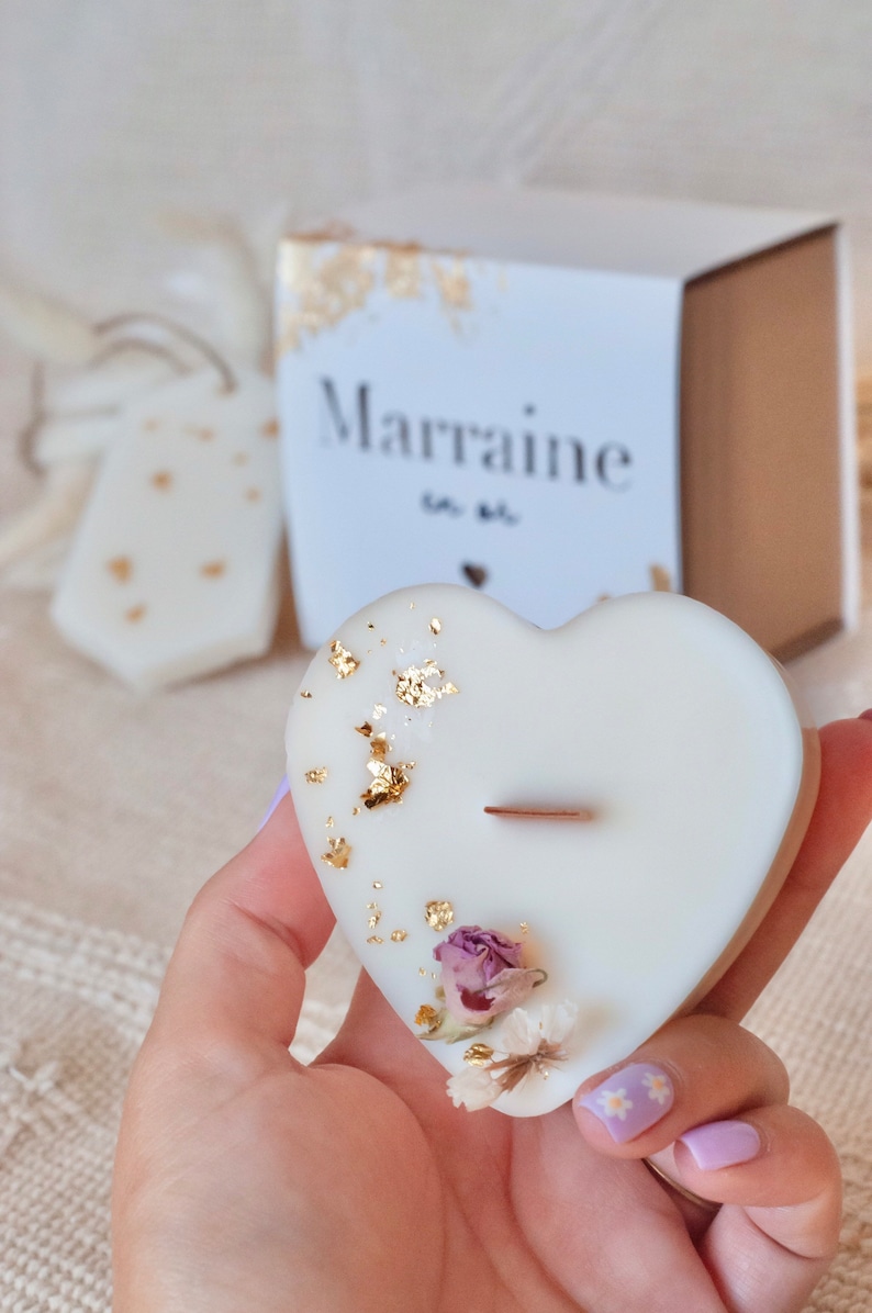 Op de afbeelding: Een hartvormige witte kaars met goudbladaccenten en gedroogde bloemen. De kaars heeft een houten lont en is versierd met kleine gedroogde bloemen. Een doos met het woord "Marraine" staat op de achtergrond.