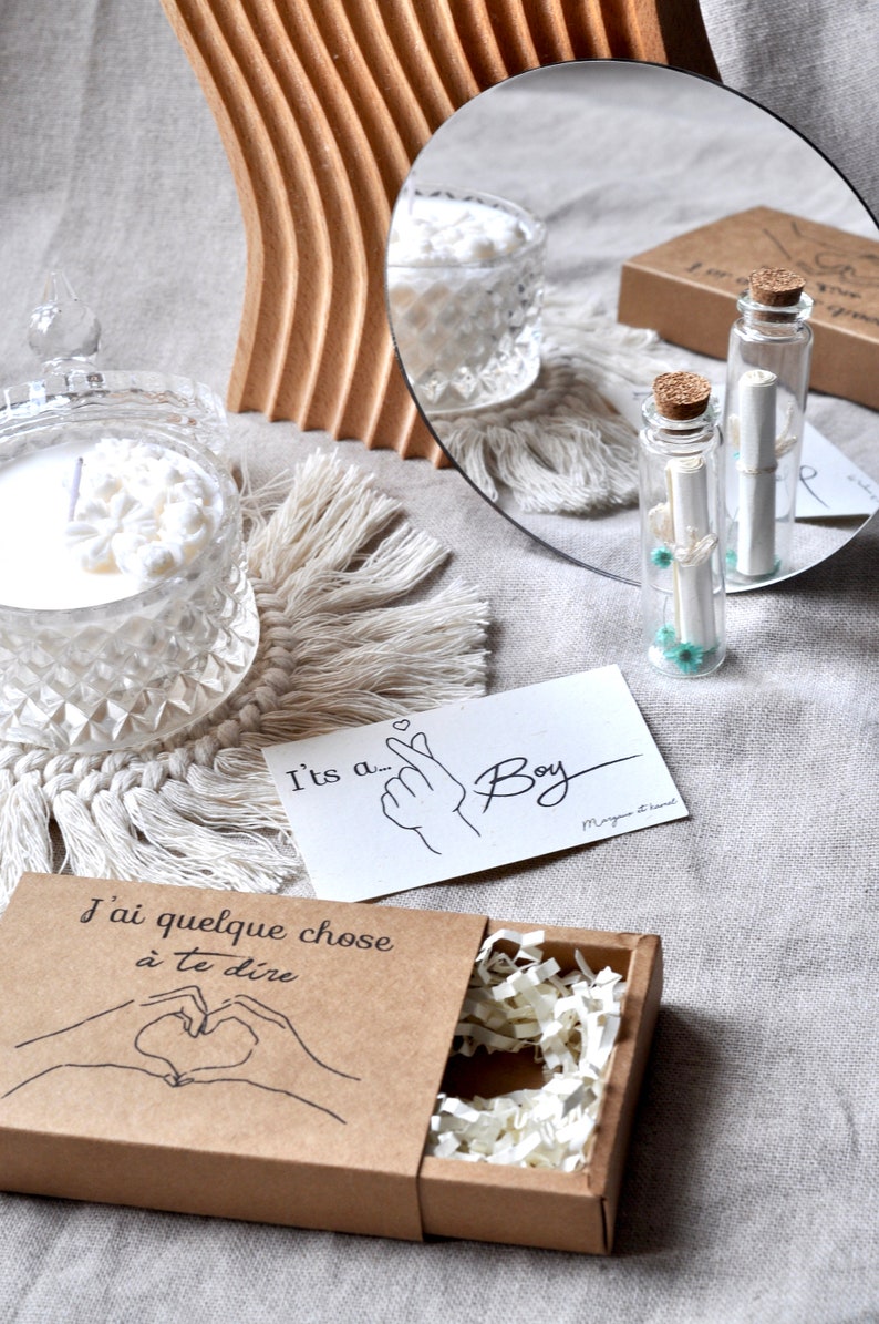 May include: A brown box with the text "J'ai quelque chose &agrave; te dire" and a hand drawn heart. The box is open and filled with white tissue paper. A white card with the text "It's a... Boy" and a hand drawn finger heart is on a white surface. A clear glass bottle with a cork stopper and a rolled up piece of paper inside is on the surface. A clear glass candle with a white wax flower on top is on a white crocheted coaster.