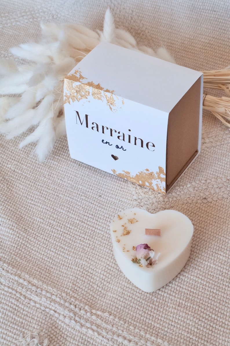 Op de afbeelding: Een witte hartvormige kaars met goudblad accenten, een houten lont en gedroogde bloemen. Een witte doos met gouden rand en de tekst "Marraine en or" is ook aanwezig.