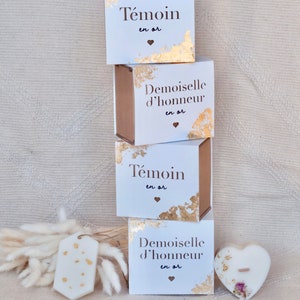 Op de afbeelding: Witte geschenkdozen met gouden folie accenten en Franse tekst. De dozen zijn gestapeld, samen met een hartvormige kaars en een label met gouden spikkels. De dozen lezen "T&eacute;moin en or" en "Demoiselle d'honneur en or".
