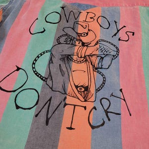 May include: A button-up shirt with vertical stripes in pink, teal, and blue. The back of the shirt features a black illustration of a cowboy and the text "Cowboys Don't Cry."