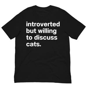Könnte beinhalten: Schwarzes T-Shirt mit dem weißen Text: "introverted but willing to discuss cats." Das Shirt hat einen Rundhalsausschnitt und kurze Ärmel. Der Text ist mittig auf der Brust platziert.