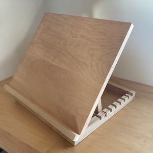 May include: A light-colored wooden easel with an angled surface for displaying artwork or holding books. The easel has a sturdy base with multiple slots for adjusting the angle. The wood grain is visible, giving it a natural look.