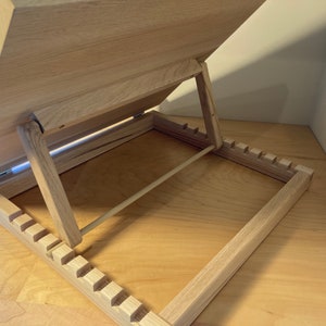 K&ouml;nnte beinhalten: Ein B&uuml;cherst&auml;nder aus Holz mit einer hellen Holzoptik. Der St&auml;nder ist ge&ouml;ffnet und die Oberseite ist nach oben geneigt. Der St&auml;nder hat eine Reihe von Schlitzen an der Unterseite, um den Winkel der Oberseite einzustellen.