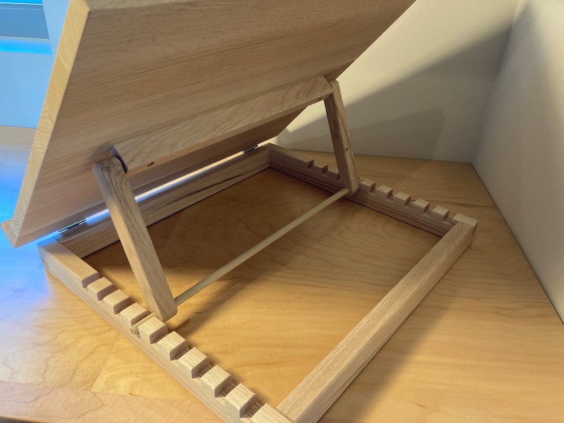 K&ouml;nnte beinhalten: Ein B&uuml;cherst&auml;nder aus Holz mit einer hellen Holzoptik. Der St&auml;nder ist ge&ouml;ffnet und die Oberseite ist nach oben geneigt. Der St&auml;nder hat eine Reihe von Schlitzen an der Unterseite, um den Winkel der Oberseite einzustellen.