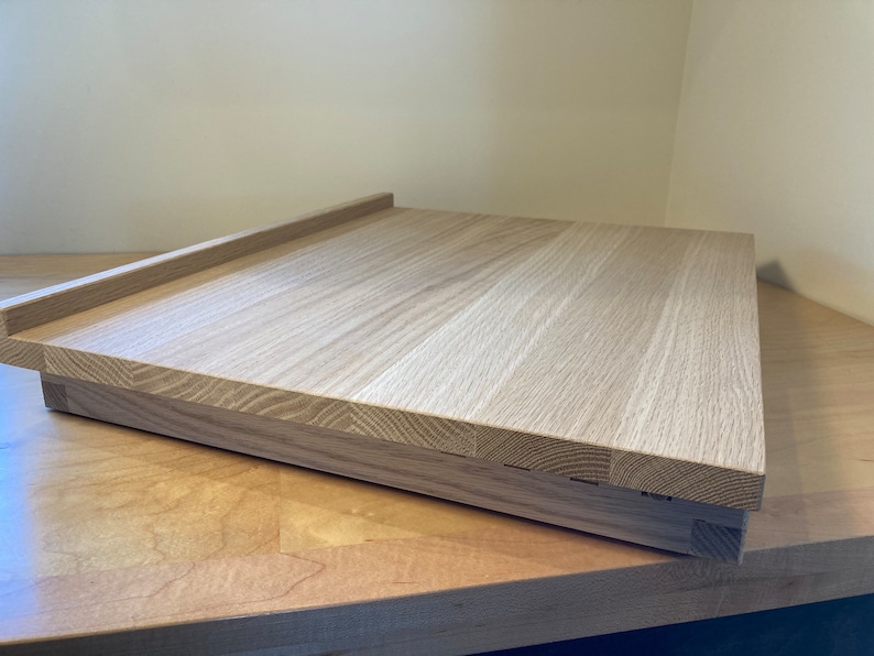 K&ouml;nnte beinhalten: Ein helles Holz-Schneidebrett mit einem erh&ouml;hten Rand auf einer Seite. Das Brett besteht aus mehreren zusammengef&uuml;gten Holzst&uuml;cken.