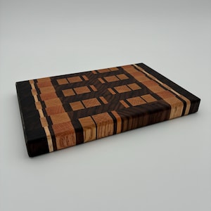 Op de afbeelding: Een rechthoekige houten snijplank met een kops houten constructie en een geometrisch patroon. Het ontwerp bestaat uit afwisselende blokken van donker walnoothout, eikenhout en licht esdoornhout, gerangschikt in een rasterpatroon.