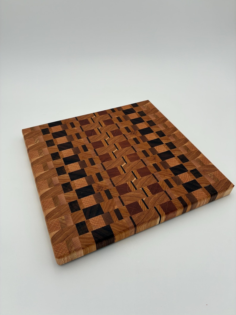 Op de afbeelding: Een vierkante houten snijplank met een kops houten constructie en een moza&iuml;ekpatroon van licht-, midden- en donkerbruine houtblokken. Het oppervlak toont een verscheidenheid aan natuurlijke houtnerven en tinten in een geometrisch raster.