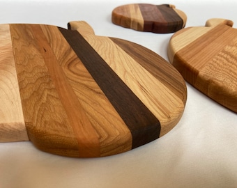 May include: Three apple-shaped cutting boards crafted from alternating vertical strips of light, medium, and dark-toned wood. Each board features a smooth, rounded surface with a small stem protrusion at the top, showcasing natural wood grain patterns.