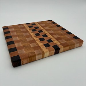 Op de afbeelding: Een vierkante houten snijplank met een kops houten constructie en een veelkleurig dambordpatroon. Het ontwerp bevat afwisselende blokken van lichtbruin, medium bruin en donker espressokleurig hout in een rasterpatroon.