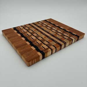 Op de afbeelding: Een rechthoekige houten snijplank met een kops houten constructie en een complex geometrisch patroon. Het oppervlak toont een moza&iuml;ek van lichte, middelbruine en donkerbruine houttinten die in afwisselende blokken zijn gerangschikt.