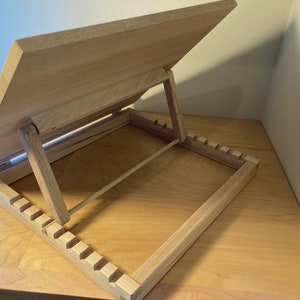K&ouml;nnte beinhalten: Ein Holzstaffelei mit einer hellen Holzoptik. Die Staffelei hat eine klappbare Oberseite, die in verschiedenen Winkeln verstellt werden kann. Die Staffelei verf&uuml;gt &uuml;ber ein integriertes Ablagefach mit einer Reihe von Schlitzen zur Aufbewahrung von Kunstbedarf.