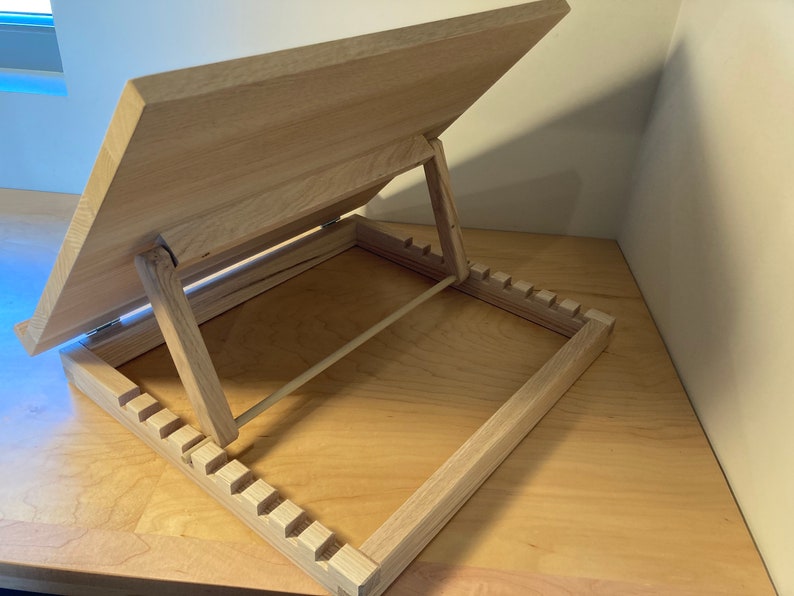 K&ouml;nnte beinhalten: Ein Holzstaffelei mit einer hellen Holzoptik. Die Staffelei hat eine klappbare Oberseite, die in verschiedenen Winkeln verstellt werden kann. Die Staffelei verf&uuml;gt &uuml;ber ein integriertes Ablagefach mit einer Reihe von Schlitzen zur Aufbewahrung von Kunstbedarf.