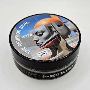 Puede incluir: Un recipiente redondo negro de jabón de manteca de mango con una etiqueta que muestra una ilustración estilizada de un perfil humano, formas geométricas naranjas y azules, y el texto THE REVISION BEHL 2026 y TALENT SOAP FACTORY en la tapa.