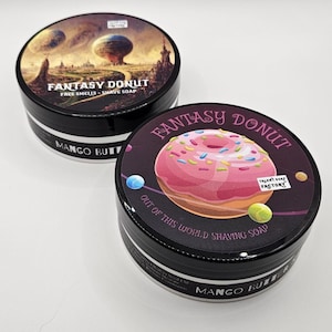 May include: Two round black containers of shaving soap labeled Fantasy Donut. One lid features a surreal landscape with planets, the other a pink frosted donut with sprinkles. Both containers have black sides with the text Mango Butter printed on them.