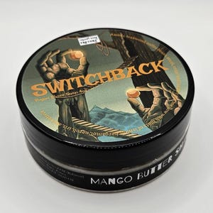 May include: A round black container of Switchback Vegan Shave Soap 4oz. The lid features a graphic illustration of two hands holding glowing orbs, a tree, and a bridge. The text reads Switchback, Vegan Shave Soap 4oz, and Mango Butter Shave.