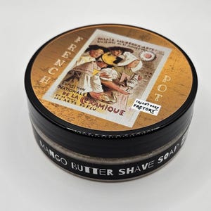 Op de afbeelding: Een ronde zwarte verpakking met Mango Butter scheerzeep. Het deksel bevat een vintage illustratie van mensen met aardewerk, de tekst "FRENCH POT", "EXPOSITION NATIONALE DE LA CERAMIQUE" en een "TALENT SOAP FACTORY" label op de zijkant.
