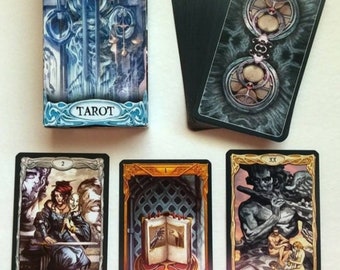 May include: A deck of Epic Tarot cards with a blue and silver box. The box has the words "Epic Tarot" on it. The cards have a black back with a silver design. The cards are shown with the 2 of Swords, the 1 of Wands, and the 20 of Judgement cards.