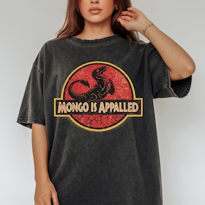 May include: A charcoal gray short-sleeve t-shirt featuring a circular graphic with a red textured background and a black dinosaur silhouette. The text "MONGO IS APPALED" is printed in a bold, light-colored font across the center of the graphic.