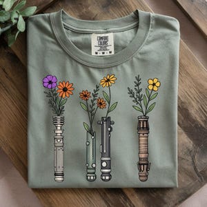 Pode incluir: Camiseta verde s&aacute;lvia com um gr&aacute;fico de tr&ecirc;s sabres de luz estilizados, cada um com uma flor diferente. As flores s&atilde;o roxas, laranja e amarelas. A camiseta tem gola redonda e uma etiqueta "Comfort Colors".