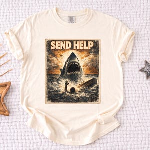 May include: Cream-colored t-shirt featuring a distressed graphic of a shark attacking a person in the ocean, with the words "SEND HELP" above the shark. The design has a vintage, weathered look.