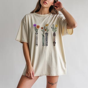 Pode incluir: Camiseta creme com um gr&aacute;fico de tr&ecirc;s sabres de luz, cada um segurando um tipo diferente de flor. As flores incluem flores roxas, laranjas e amarelas. O desenho &eacute; centrado na frente da camiseta.