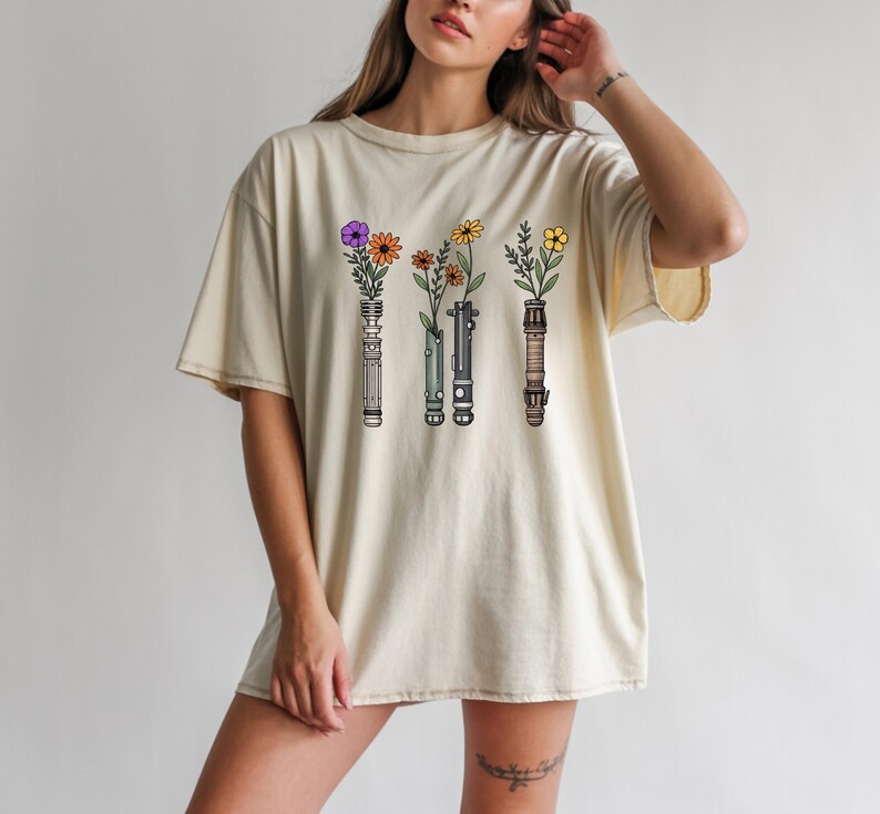 Pode incluir: Camiseta creme com um gr&aacute;fico de tr&ecirc;s sabres de luz, cada um segurando um tipo diferente de flor. As flores incluem flores roxas, laranjas e amarelas. O desenho &eacute; centrado na frente da camiseta.