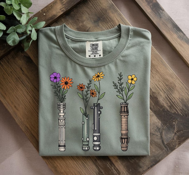 Pode incluir: Camiseta verde s&aacute;lvia com um gr&aacute;fico de tr&ecirc;s sabres de luz estilizados, cada um com uma flor diferente. As flores s&atilde;o roxas, laranja e amarelas. A camiseta tem gola redonda e uma etiqueta "Comfort Colors".