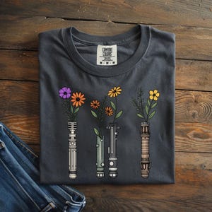 Pode incluir: Camiseta cinza escuro com um design floral. O design inclui ilustra&ccedil;&otilde;es de sabres de luz com flores e vegeta&ccedil;&atilde;o coloridas. A camiseta tem gola redonda e uma etiqueta que diz "Comfort Colors".