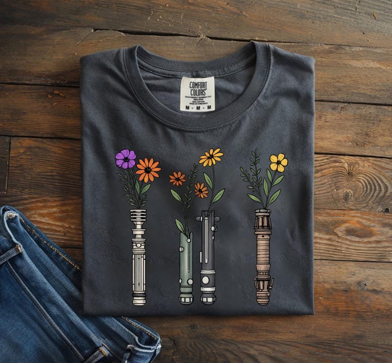 Pode incluir: Camiseta cinza escuro com um design floral. O design inclui ilustra&ccedil;&otilde;es de sabres de luz com flores e vegeta&ccedil;&atilde;o coloridas. A camiseta tem gola redonda e uma etiqueta que diz "Comfort Colors".