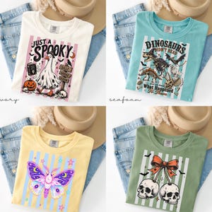 May include: Four crew neck t-shirts in ivory, seafoam, yellow, and sage green. The ivory shirt features a ghost goose and text 'Just a Spooky'. The seafoam shirt shows dinosaur skeletons and text 'Dinosaurs didn't read'. The yellow shirt displays a moth, and the sage green shirt shows two skulls with a bow.