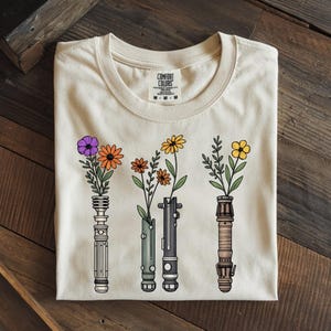 Pode incluir: Camiseta bege com um design floral. O gr&aacute;fico apresenta quatro sabres de luz estilizados, cada um segurando uma flor diferente. As flores s&atilde;o roxas, laranja, amarelas e amarelas. A camisa &eacute; da Comfort Colors.