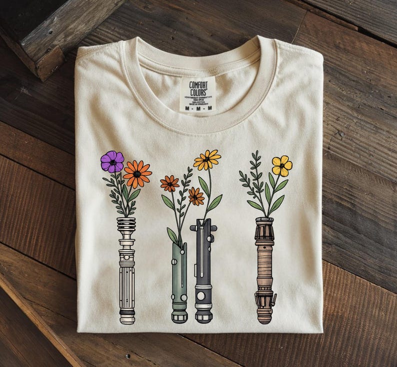 Pode incluir: Camiseta bege com um design floral. O gr&aacute;fico apresenta quatro sabres de luz estilizados, cada um segurando uma flor diferente. As flores s&atilde;o roxas, laranja, amarelas e amarelas. A camisa &eacute; da Comfort Colors.