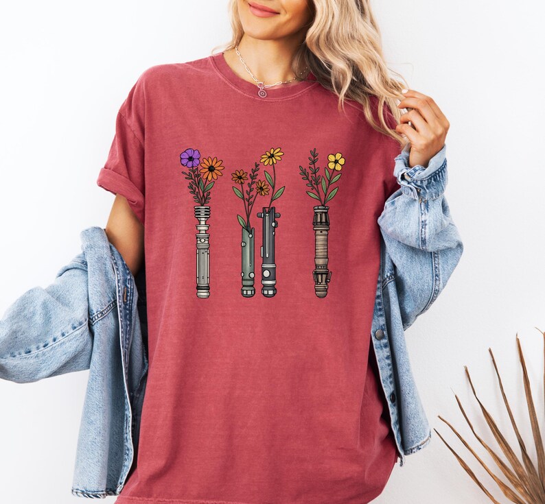 Pode incluir: Camiseta vermelho-alaranjada com um desenho de tr&ecirc;s sabres de luz, cada um segurando uma flor diferente. Os sabres de luz s&atilde;o em tons de prata, cinza e marrom, com flores roxas, amarelas e laranja. Estilo casual.