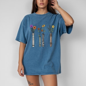 Pode incluir: Camiseta azul com um desenho de tr&ecirc;s cabos de sabre de luz, cada um com uma flor diferente. As flores s&atilde;o roxas, laranjas e amarelas. A camiseta tem um corte folgado e mangas curtas.