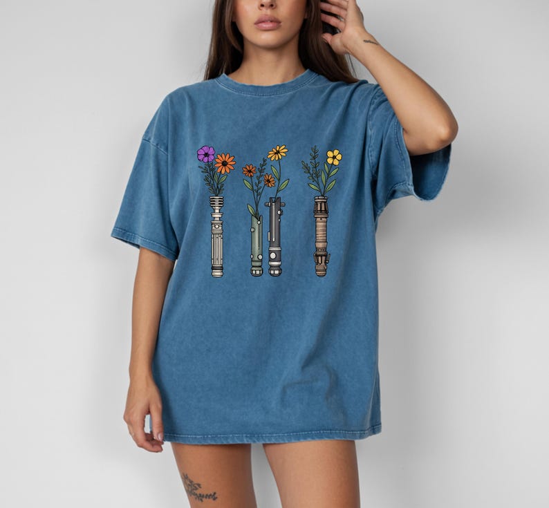 Pode incluir: Camiseta azul com um desenho de tr&ecirc;s cabos de sabre de luz, cada um com uma flor diferente. As flores s&atilde;o roxas, laranjas e amarelas. A camiseta tem um corte folgado e mangas curtas.