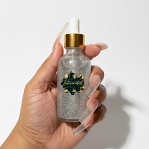 May include: A small, clear glass bottle filled with silver glitter, held against a white backdrop. The bottle has a gold cap and a white dropper. The label reads "Intoxicated" in a stylized font.