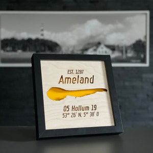 May include: Wooden framed artwork of Ameland island, established in 1287. The island's shape is cut out, revealing a yellow background. Includes the address 05 Hollum 19 and coordinates 53° 26' N, 5° 38' 0. A black and white photo hangs in the background.
