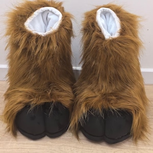 May include: A pair of novelty brown furry slippers shaped like animal feet. The slippers have a black sole and a white interior. The faux fur is a light brown color.