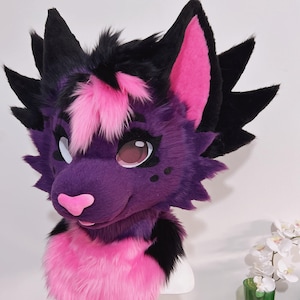 May include: A purple and pink furry fursuit head with black accents. The head features large, expressive eyes, a pink nose, and a smiling mouth. The ears are lined with pink, and the fur is a mix of purple, pink, and black.