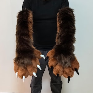 May include: A pair of brown furry hand paws with white claws. The paws are being worn by a person, but the focus is on the paws themselves.
