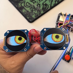 Peut inclure: Deux circuits imprimés ronds bleus avec des yeux jaunes et orange imprimés dessus. Les cartes sont reliées par des fils rouges.