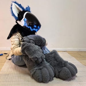 May include: A furry fursuit head with blue and white accents, a black visor, and glowing blue lights. The suit includes a black fur collar, a tan jacket, and large gray furry paws. The person is sitting on a beige mat.