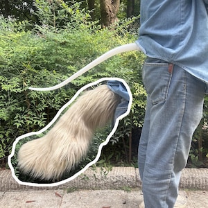 Peut inclure: Une longue queue en fausse fourrure beige fixée sur une base flexible blanche. La queue est portée à la taille, s'étendant depuis un jean bleu et une chemise en jean bleu clair, avec une texture épaisse et hirsute et une extrémité effilée.