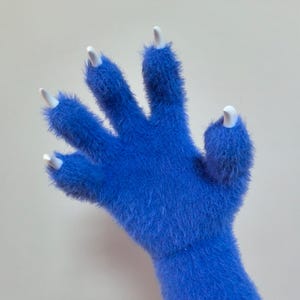May include: A blue fuzzy hand with white claws. The hand is raised with the fingers spread apart.