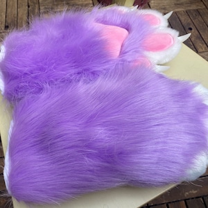 May include: A pair of fluffy purple and white paw-shaped hand warmers. The warmers are made of soft, plush fabric and have a white trim around the edges.
