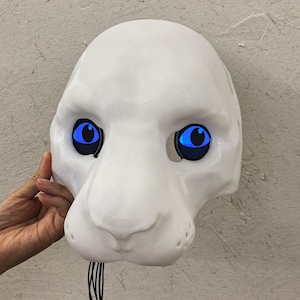 Può includere: Maschera di animale bianca con occhi blu. La maschera è realizzata in un materiale bianco liscio e ha un viso di animale realistico.