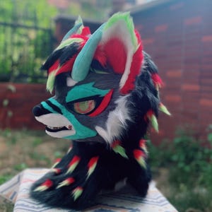 Puede incluir: Una cabeza de animal peluda de colores con pelo negro, blanco, rojo, verde y azul. La cabeza tiene un hocico largo y dientes afilados. Los ojos son negros con un patrón de espiral rojo y verde.